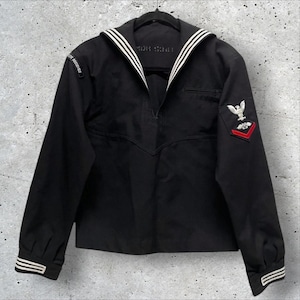 May include: Black vintage sailor shirt with white stripes on the collar and cuffs. Features a small pocket and patches on the sleeves. The shirt is hanging on a black hanger.