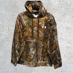 May include: A long-sleeve hooded sweatshirt featuring a brown, green, and tan forest camouflage pattern. The garment includes a front kangaroo pocket, a drawstring hood, and a white Under Armour logo printed on the upper left chest area.