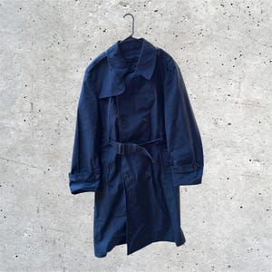 May include: A dark navy blue double-breasted trench coat featuring a wide collar, shoulder epaulets, long sleeves with buttoned cuffs, and a matching fabric belt with a rectangular buckle at the waist, all hanging on a black plastic hanger.