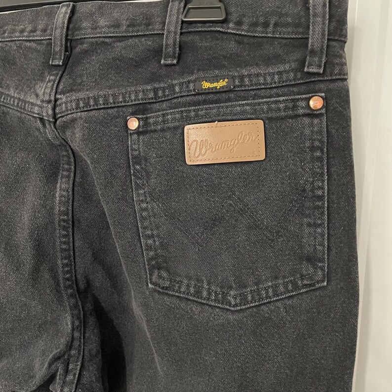 May include: Close-up of the back of black Wrangler jeans. The jeans have a classic five-pocket design with a brown leather patch on the back pocket. The Wrangler logo is visible on the patch and above the back pocket.