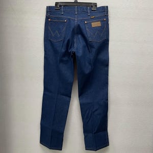 May include: Dark blue denim jeans with the Wrangler logo on the back pockets and a brown leather patch. The jeans are hanging on a black hanger. The jeans have copper-colored rivets and stitching.