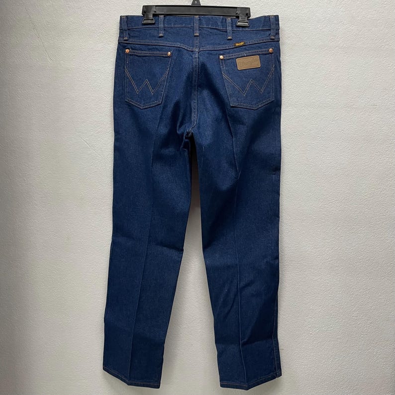 May include: Dark blue denim jeans with the Wrangler logo on the back pockets and a brown leather patch. The jeans are hanging on a black hanger. The jeans have copper-colored rivets and stitching.