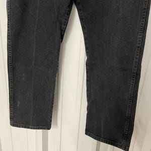 May include: Close-up of black denim jeans. The jeans have a straight leg cut and are made of a dark wash denim material. The stitching is visible along the seams and pockets. The jeans are slightly faded in areas, giving them a worn look.