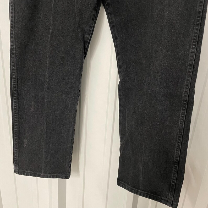 May include: Close-up of black denim jeans. The jeans have a straight leg cut and are made of a dark wash denim material. The stitching is visible along the seams and pockets. The jeans are slightly faded in areas, giving them a worn look.
