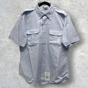 May include: A light blue short-sleeve shirt with two chest pockets and epaulets. The shirt has a button-down front and a collar. A tag is visible near the bottom. The shirt is hanging on a black hanger.