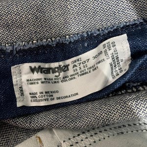 May include: Close-up of a Wrangler jeans label. The label is white with black text, including the brand name, size 35 x 32, and care instructions. The jeans are a dark blue denim.