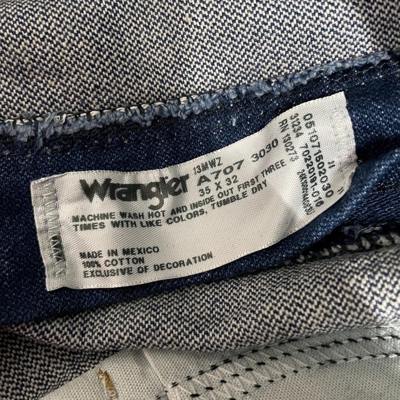 May include: Close-up of a Wrangler jeans label. The label is white with black text, including the brand name, size 35 x 32, and care instructions. The jeans are a dark blue denim.