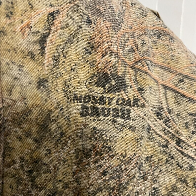 May include: Close-up of a camouflage fabric with the text "Mossy Oak Brush" in dark green. The fabric has a pattern of brown, tan, and green, creating a natural, outdoor aesthetic. The fabric appears to be a durable material.