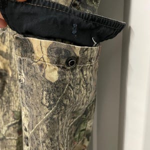 May include: Close-up of camouflage fabric with a black pocket lining and a metal snap closure. The fabric features a pattern of green, brown, and tan, typical of hunting or outdoor apparel. The pocket is partially open, revealing the interior.
