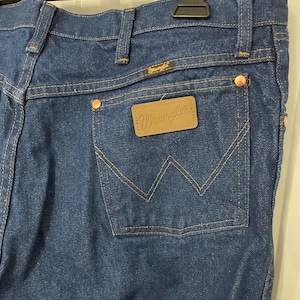 May include: Close-up of dark blue denim jeans with a classic five-pocket design. Features include copper rivets, a brown leather patch with the "Wrangler" logo, and the Wrangler label above the back pocket. The stitching is a contrasting tan color.