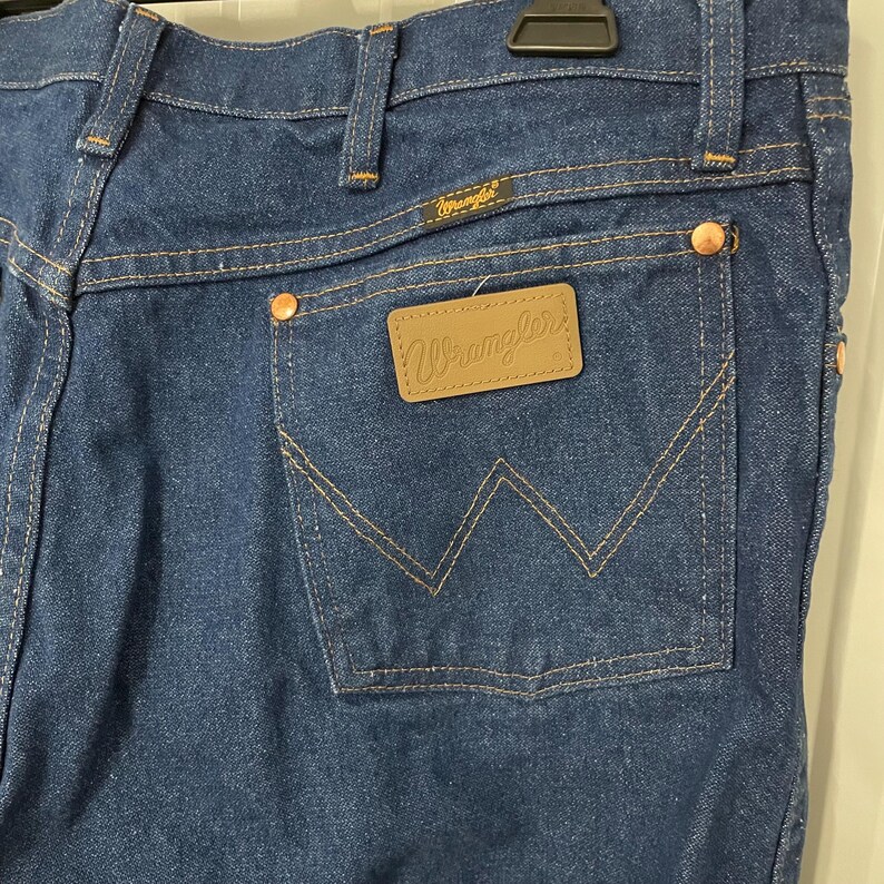 May include: Close-up of dark blue denim jeans with a classic five-pocket design. Features include copper rivets, a brown leather patch with the "Wrangler" logo, and the Wrangler label above the back pocket. The stitching is a contrasting tan color.
