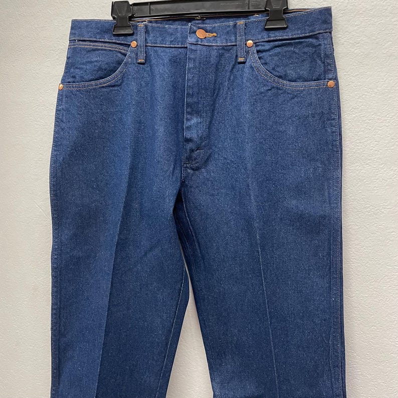 May include: Close-up of a pair of blue denim jeans. The jeans have a classic five-pocket design, with copper rivets and a button closure. The denim has a medium wash and a straight leg cut.