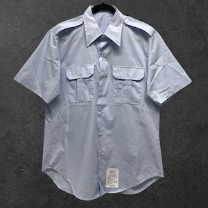 Puede incluir: Una camisa azul claro de manga corta con dos bolsillos en el pecho y charreteras. La camisa tiene una botonadura frontal y un cuello. Una pequeña etiqueta está cosida en la parte inferior. La camisa está colgada de una percha negra.