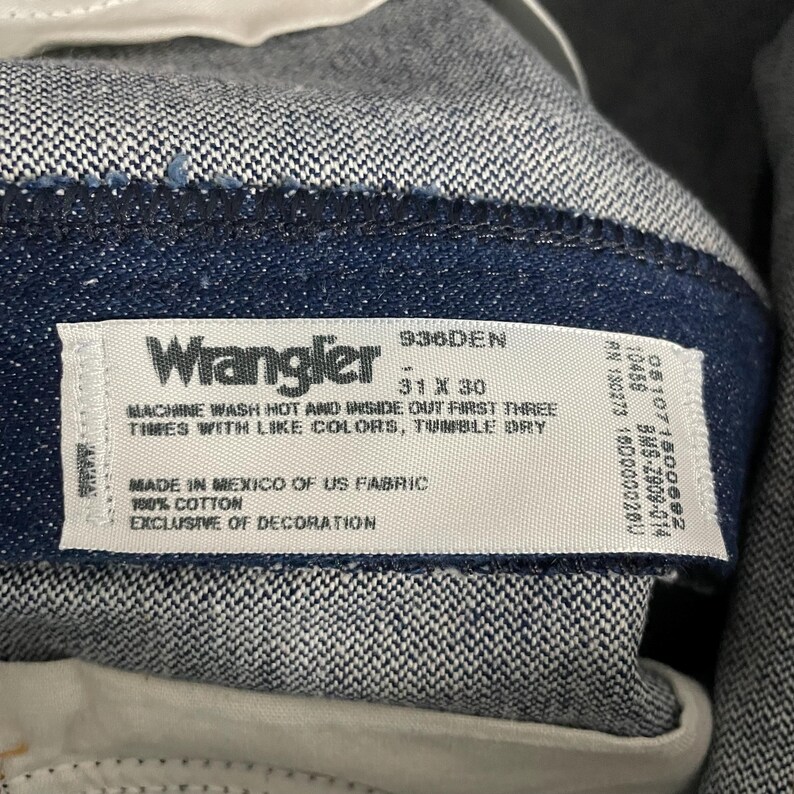 Puede incluir: Primer plano de unos vaqueros Wrangler de mezclilla. La etiqueta dice "Wrangler 31 x 30" con instrucciones de lavado. Los vaqueros son 100% algod&oacute;n y est&aacute;n fabricados en M&eacute;xico.