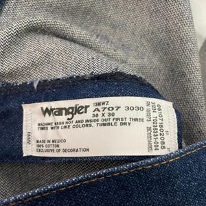 Op de afbeelding: Close-up van een Wrangler jeanslabel. Het witte label heeft zwarte tekst, inclusief de merknaam, het modelnummer en de maat 36 x 30. Het label vermeldt ook dat de jeans gemaakt zijn van 100% katoen en gemaakt in Mexico.