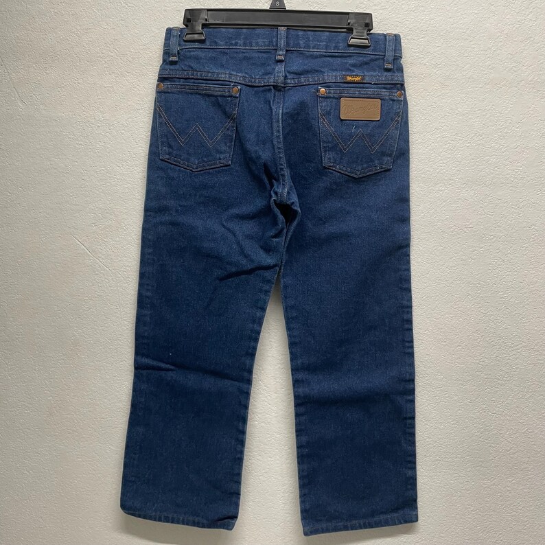 May include: Dark blue denim jeans with a classic five-pocket design. Features include a brown leather patch and the Wrangler logo. The jeans are hanging on a black hanger.