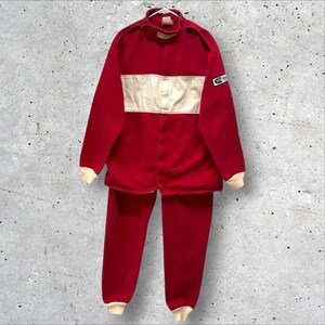 May include: A red racing suit with a cream-colored horizontal stripe across the chest and cream-colored cuffs. The suit includes a jacket and matching pants. A small patch is visible on the right sleeve. The suit is displayed against a mottled gray background.