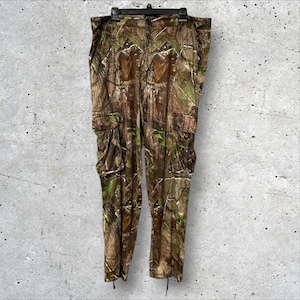 Puede incluir: Pantalones cargo de camuflaje con múltiples bolsillos, colgados de una percha negra. Los pantalones presentan un patrón de camuflaje de bosque marrón, verde y beige. Los pantalones están hechos de un material ligero.