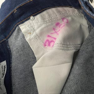 Puede incluir: Primer plano del interior de unos vaqueros azules. El forro blanco del bolsillo tiene texto rosa que dice "31x30". Los vaqueros tienen cierre de bot&oacute;n y una costura visible.