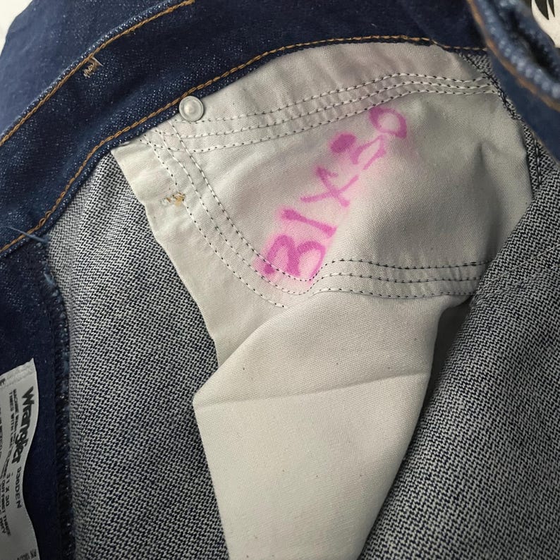 Puede incluir: Primer plano del interior de unos vaqueros azules. El forro blanco del bolsillo tiene texto rosa que dice "31x30". Los vaqueros tienen cierre de bot&oacute;n y una costura visible.