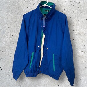 May include: A vintage blue windbreaker jacket with a high collar and a zipper closure. The jacket has green and white accents on the front and a drawstring at the bottom. The jacket is hanging on a white hanger.