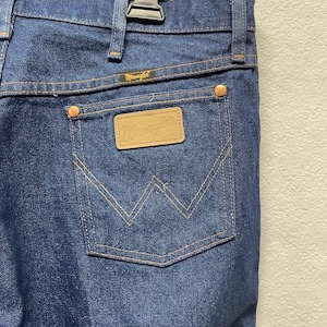 Op de afbeelding: Close-up van de achterzak van een blauwe Wrangler-jeans. De zak heeft een gestikt ontwerp en een kleine, rechthoekige leren patch met het Wrangler-logo. De jeans heeft koperkleurige klinknagels en een metalen ritssluiting.