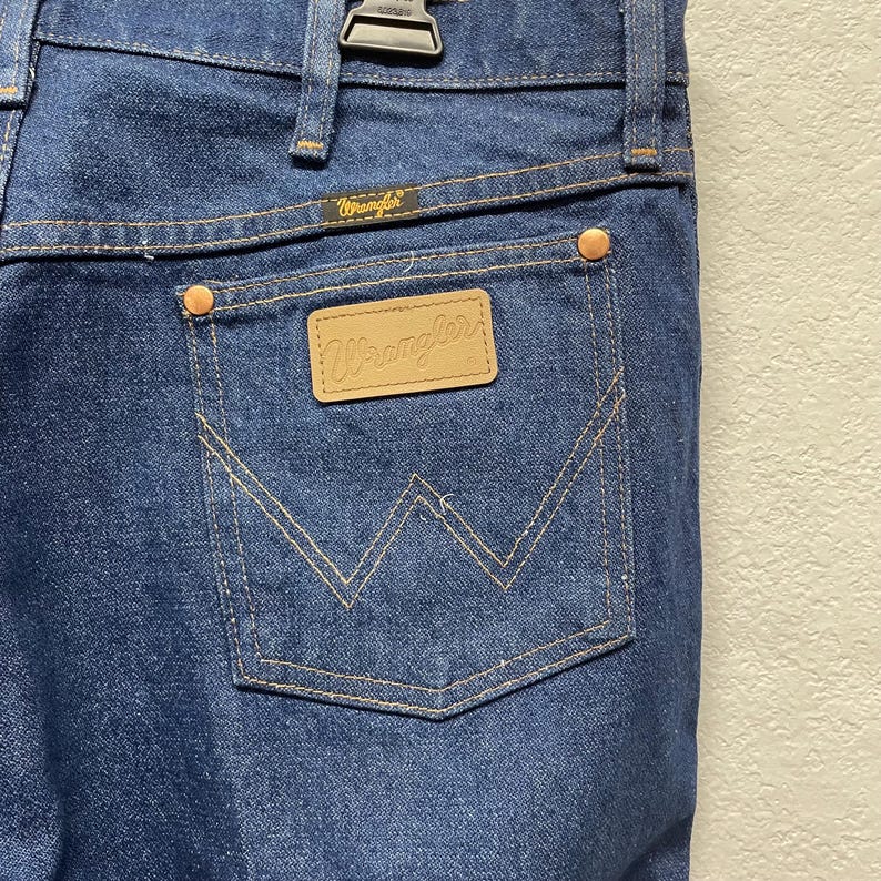 Op de afbeelding: Close-up van de achterzak van een blauwe Wrangler-jeans. De zak heeft een gestikt ontwerp en een kleine, rechthoekige leren patch met het Wrangler-logo. De jeans heeft koperkleurige klinknagels en een metalen ritssluiting.