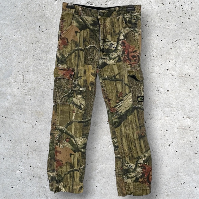 May include: Camouflage cargo pants with a button and zipper closure. The pants feature multiple pockets and a woodland camouflage pattern in shades of green, brown, and tan. The pants are hanging on a black hanger.