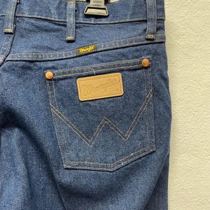 May include: Close-up of the back of blue denim Wrangler jeans. The jeans have a brown leather patch with the Wrangler logo on the back pocket. The pocket is outlined with tan stitching. The jeans have copper rivets and a Wrangler label above the back pocket.