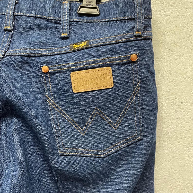May include: Close-up of the back of blue denim Wrangler jeans. The jeans have a brown leather patch with the Wrangler logo on the back pocket. The pocket is outlined with tan stitching. The jeans have copper rivets and a Wrangler label above the back pocket.