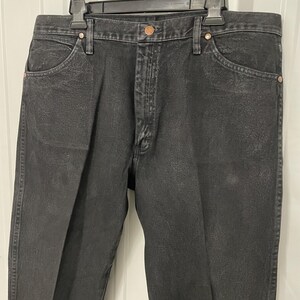May include: Black denim jeans with a classic five-pocket design. The jeans have a button and zipper closure, belt loops, and copper-colored rivets. The fabric appears slightly faded, giving them a worn look. The jeans are hanging on a black hanger.