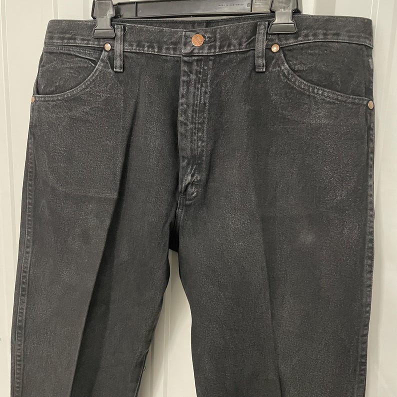May include: Black denim jeans with a classic five-pocket design. The jeans have a button and zipper closure, belt loops, and copper-colored rivets. The fabric appears slightly faded, giving them a worn look. The jeans are hanging on a black hanger.