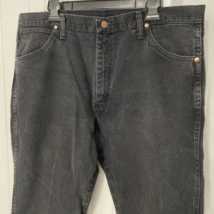 May include: Black denim jeans with a classic five-pocket design. The jeans feature copper-colored rivets and a button closure. The jeans are hanging on a black hanger.