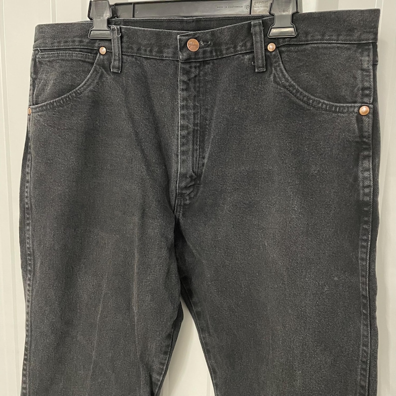 May include: Black denim jeans with a classic five-pocket design. The jeans feature copper-colored rivets and a button closure. The jeans are hanging on a black hanger.