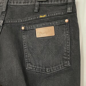May include: Close-up of black denim jeans with a Wrangler label on the back pocket. The jeans have a classic five-pocket design, with copper-colored rivets and a Wrangler logo above the back pocket.