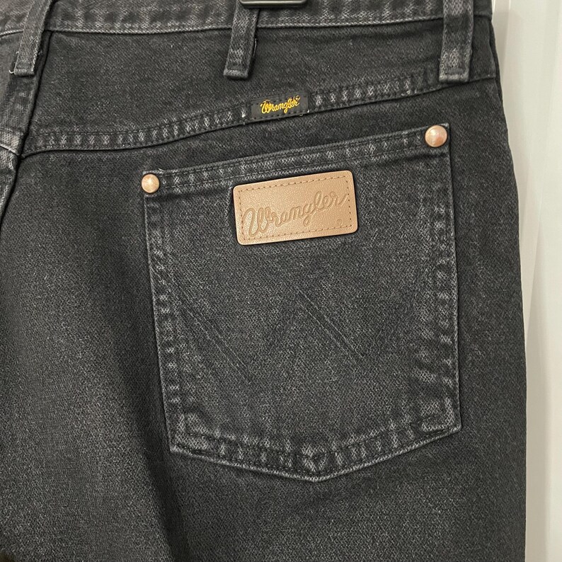 May include: Close-up of black denim jeans with a Wrangler label on the back pocket. The jeans have a classic five-pocket design, with copper-colored rivets and a Wrangler logo above the back pocket.