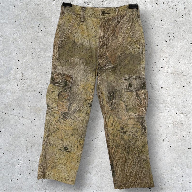 May include: Camouflage cargo pants in shades of brown, green, and tan. The pants feature multiple pockets, a button and zipper closure, and belt loops. The fabric has a textured appearance, and the pants are designed for outdoor activities.