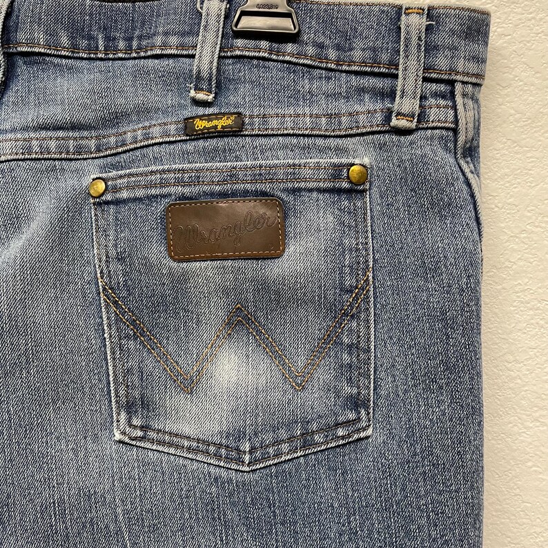 Może przedstawiać: Zbliżenie jasnoniebieskich dżins&oacute;w. Tylna kieszeń ma brązową sk&oacute;rzaną naszywkę ze złotym wytłoczonym napisem "Wrangler". Kieszeń jest przeszyta charakterystycznym wzorem "W". Dżinsy mają metalowy guzik i etykietę "Wrangler".