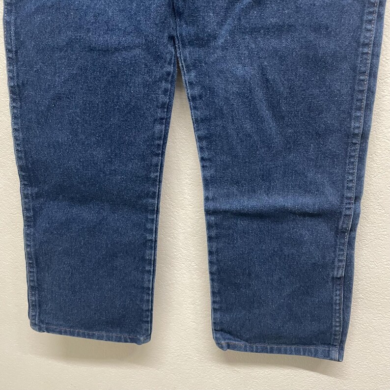 May include: Close-up of blue denim jeans. The jeans have a classic five-pocket design, with a straight leg cut. The denim fabric is a medium wash, showing subtle variations in color.