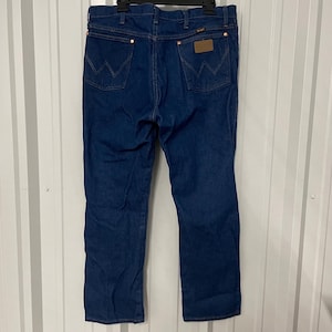 May include: Dark blue denim jeans with a classic five-pocket design. The jeans feature copper-colored rivets and a brown leather patch on the back. The Wrangler logo is visible on the back pockets.