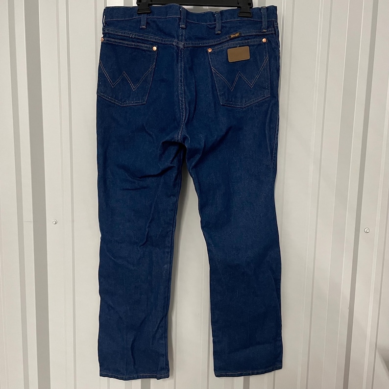 May include: Dark blue denim jeans with a classic five-pocket design. The jeans feature copper-colored rivets and a brown leather patch on the back. The Wrangler logo is visible on the back pockets.