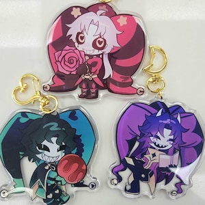 Puede incluir: Tres llaveros acrílicos con personajes de dibujos animados con rasgos exagerados. Los llaveros tienen herrajes dorados. Un personaje tiene detalles en rosa y rojo, otro en verde azulado y negro, y el último en morado y negro.