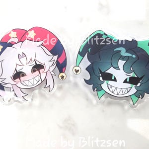 May include: Two acrylic charms featuring stylised cartoon characters with sharp teeth and jester hats. The left charm has white hair and a red and blue hat with yellow stars. The right charm has dark hair and a green and black hat with heart icons.