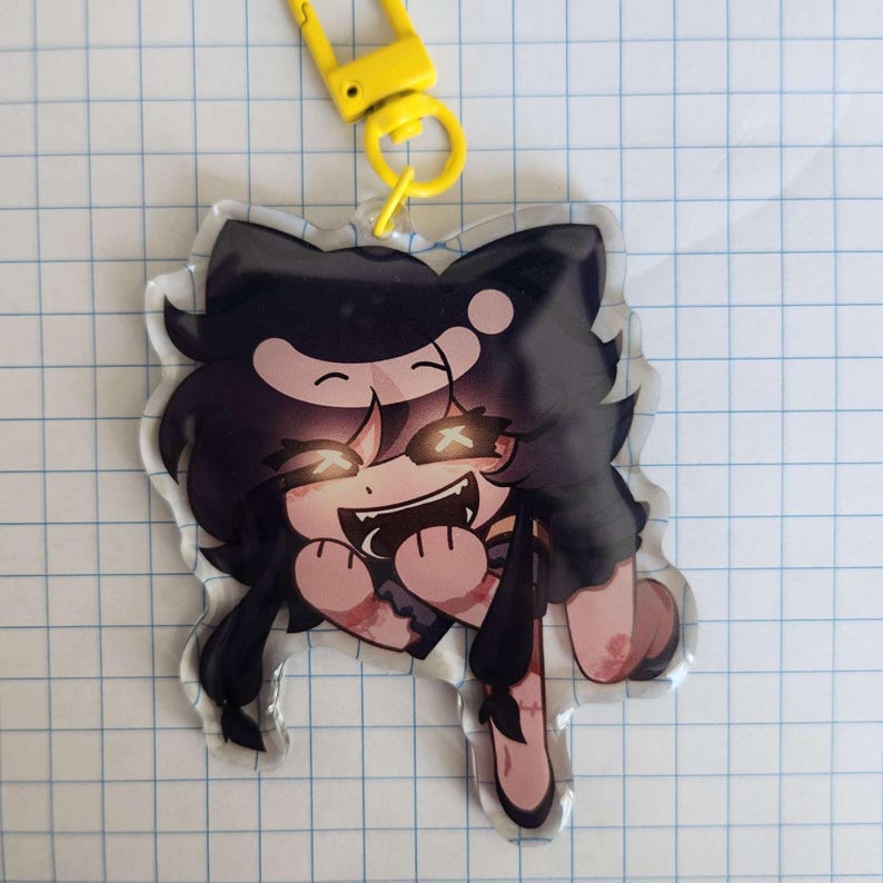 May include: A clear acrylic charm featuring a cartoon character with black hair, glowing eyes, and a wide grin. The charm has a yellow clasp and a split ring. The character has a cat-like appearance.