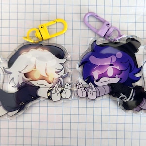 May include: Two acrylic keychains featuring anime-style characters. One has white hair and a yellow clasp, the other has purple hair and a purple clasp. Both characters are in a lying down pose with their hands up.