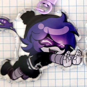 May include: A clear acrylic charm featuring a cartoon character with purple hair and glowing eyes. The character is wearing a black outfit with gray accents and is attached to a purple clasp. The charm is designed with a cartoon style.