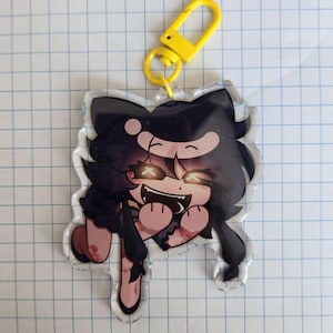 May include: A clear acrylic charm featuring a cartoon character with black hair and glowing eyes. The character has a wide grin and is holding its arms out. A yellow clasp is attached to the top.