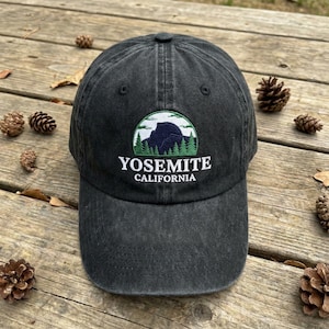 May include: A charcoal grey baseball cap featuring an embroidered patch on the front. The patch displays a dark blue mountain silhouette against a white sky, framed by green pine trees, with the text YOSEMITE CALIFORNIA printed below.
