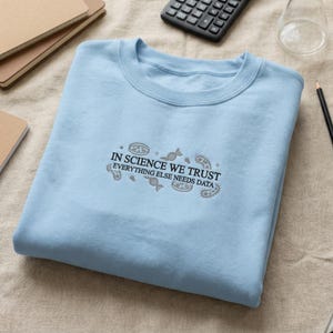 May include: A pale blue sweatshirt featuring the text "IN SCIENCE WE TRUST EVERYTHING ELSE NEEDS DATA" in grey, with scientific illustrations. The garment is made of a soft material and is suitable for casual wear.