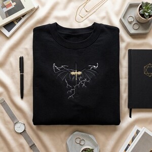 May include: A black crewneck sweatshirt featuring a central embroidered design of a gold dragon with spread wings, positioned vertically over a sword, surrounded by white lightning bolt patterns stitched directly onto the fabric.